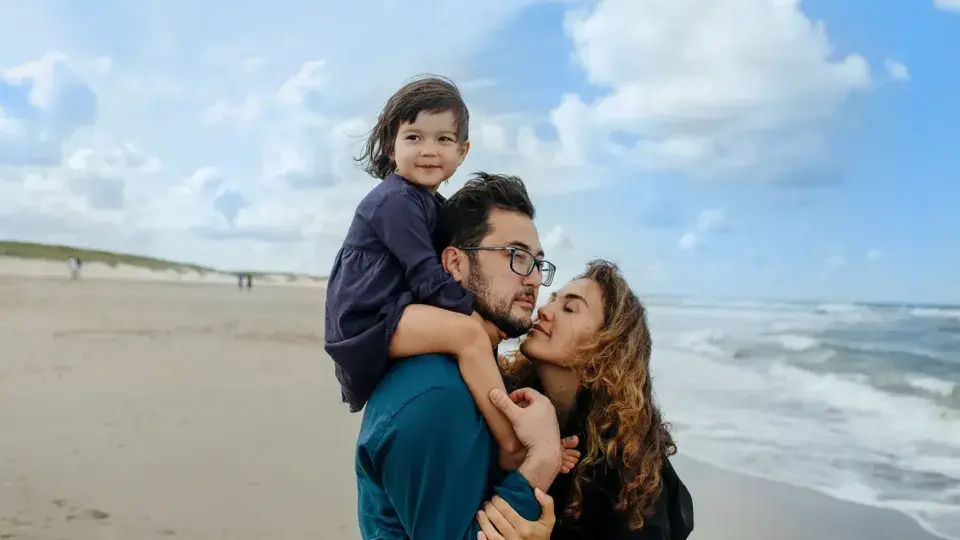 A joyful family enjoys a sunny beach day together. The father, wearing glasses and a broad smile, carries his young daughter on his shoulders, her laughter echoing across the shore. The mother walks alongside them, her face glowing with contentment as she watches her family play. The little girl, her eyes sparkling with excitement, clings onto her father's head with tiny hands, relishing the adventure. The sound of crashing waves provides a soothing backdrop to their happiness, as they create cherished memo