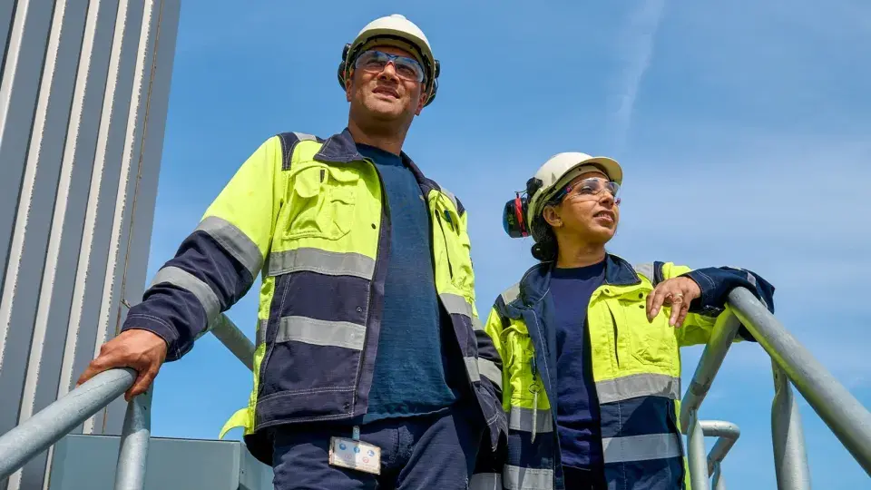 Two industrial workers, a man and a woman, stand on a metal platform in protective gear, including hard hats, reflective jackets, and safety glasses. They look out into the distance under a clear blue sky, suggesting a focus on future goals or ongoing work at an industrial site. The image highlights teamwork and safety in an outdoor work environment.
