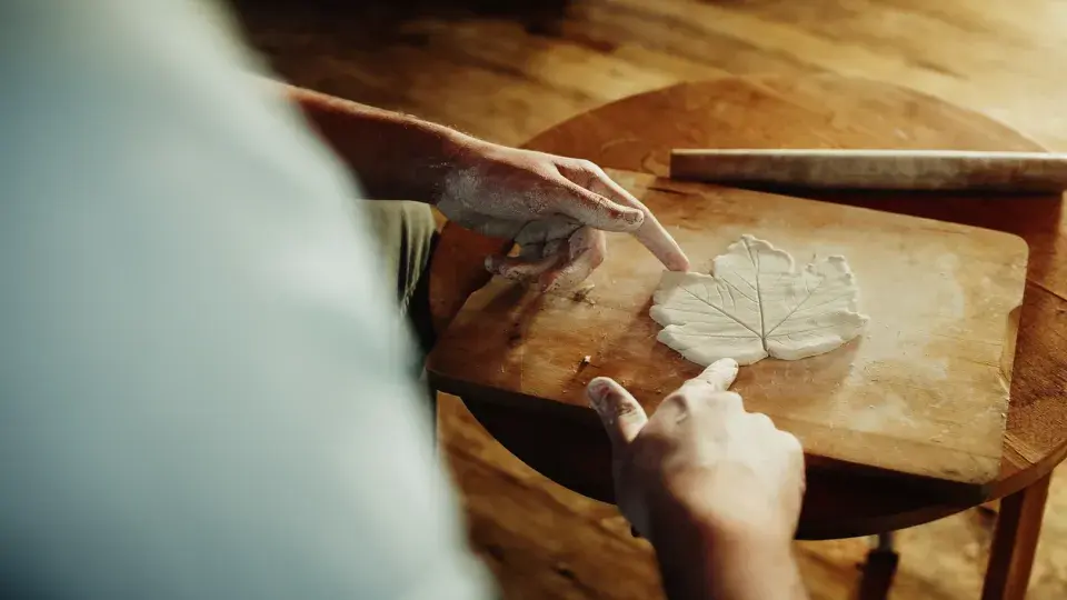 A person with clay-covered hands shapes a leaf pattern out of clay on a wooden board. The scene captures a moment of careful craftsmanship and creativity in a warm, natural setting.