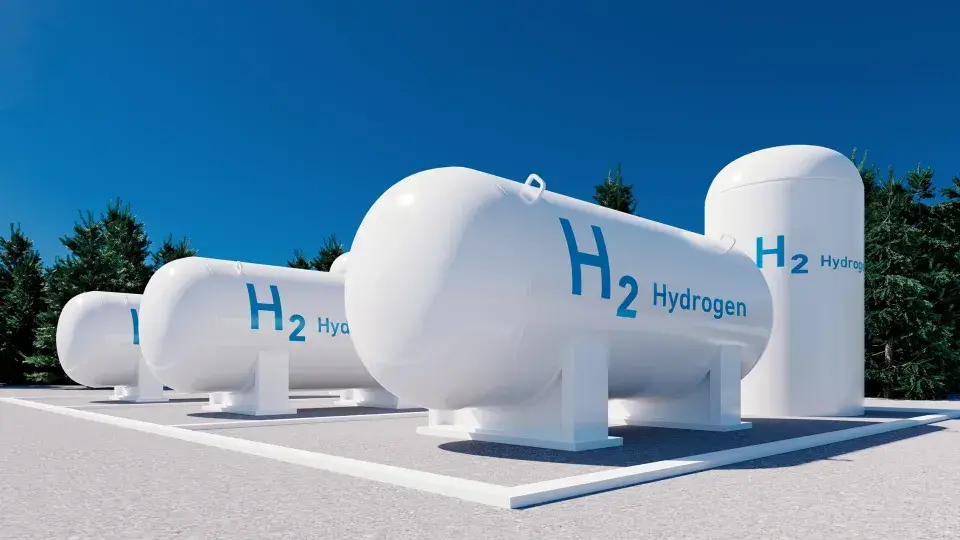 White hydrogen storage tanks labeled "H2 Hydrogen" against a clear blue sky and surrounded by trees, representing green hydrogen technology.