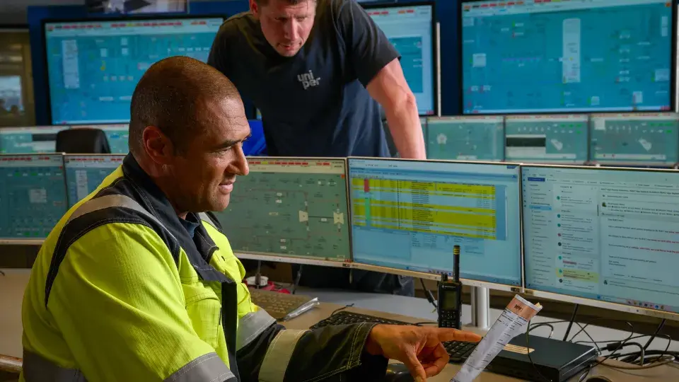Two Uniper employees working in a control room with multiple monitors displaying process data; one employee in high-visibility clothing points at a document while the other observes.