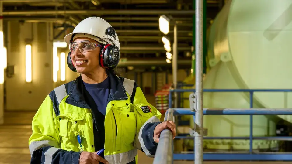 Worker wearing a yellow safety jacket, helmet, ear protection, and safety glasses, standing inside an industrial facility and looking to the side with a smile